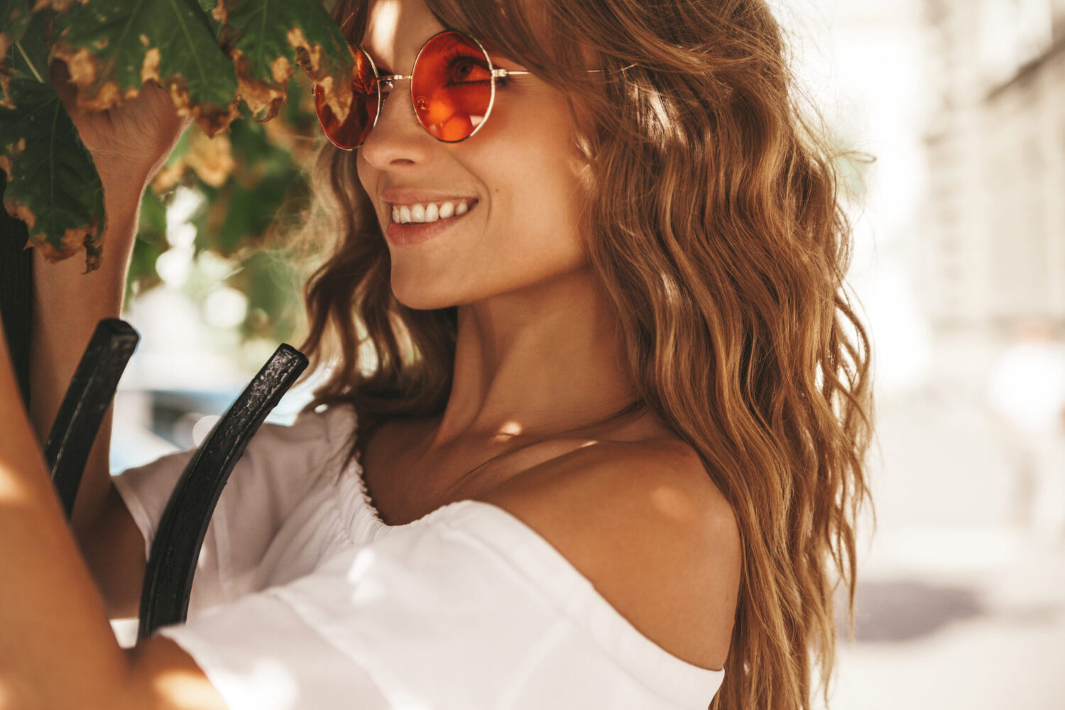 Retrato de una hermosa y linda modelo adolescente rubia sin maquillaje en ropa de vestir blanca hipster de verano posando en el fondo de la calle con gafas de sol tocando hojas de árboles